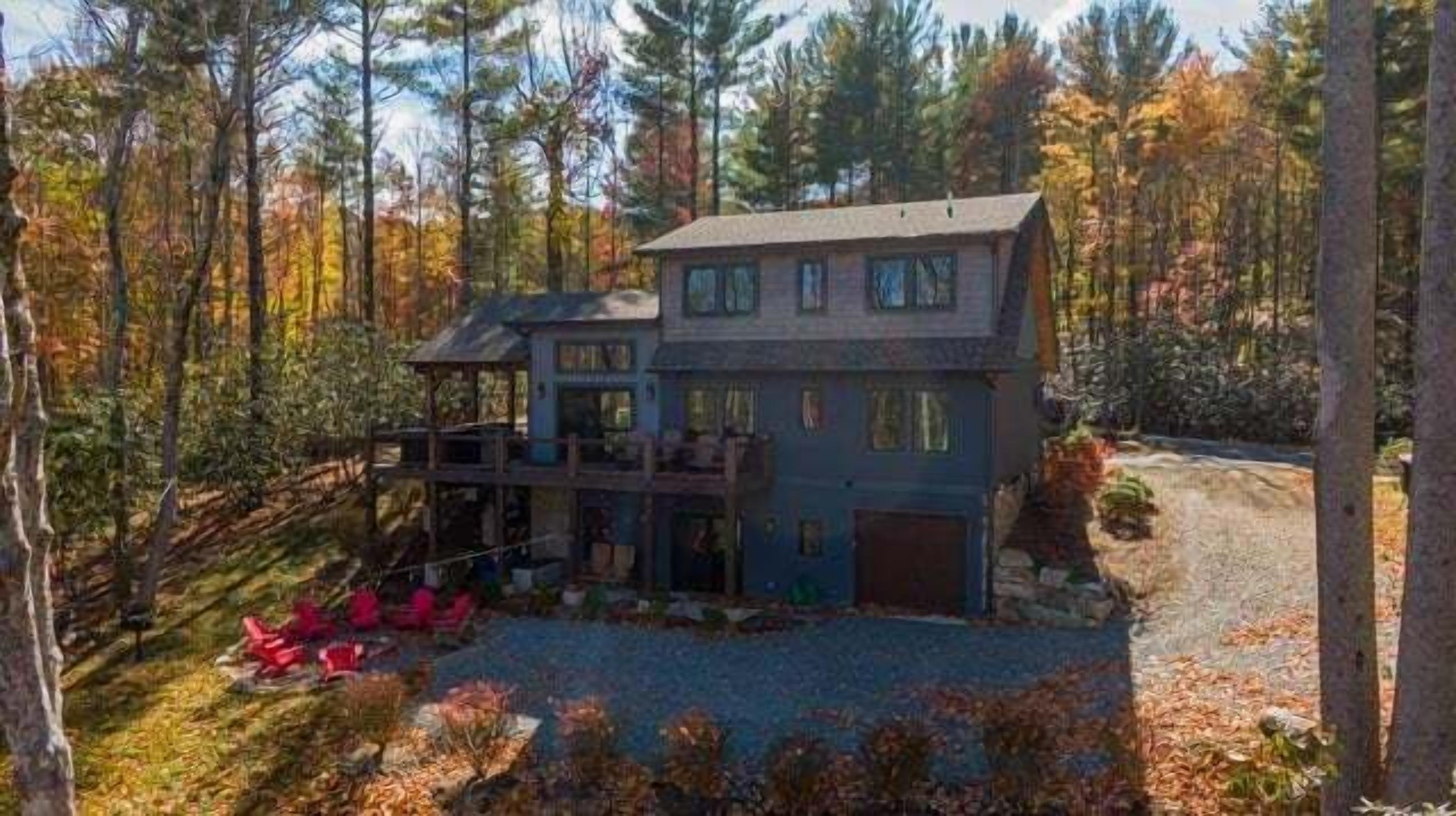A two-story house with a deck, surrounded by trees and a gravel driveway.