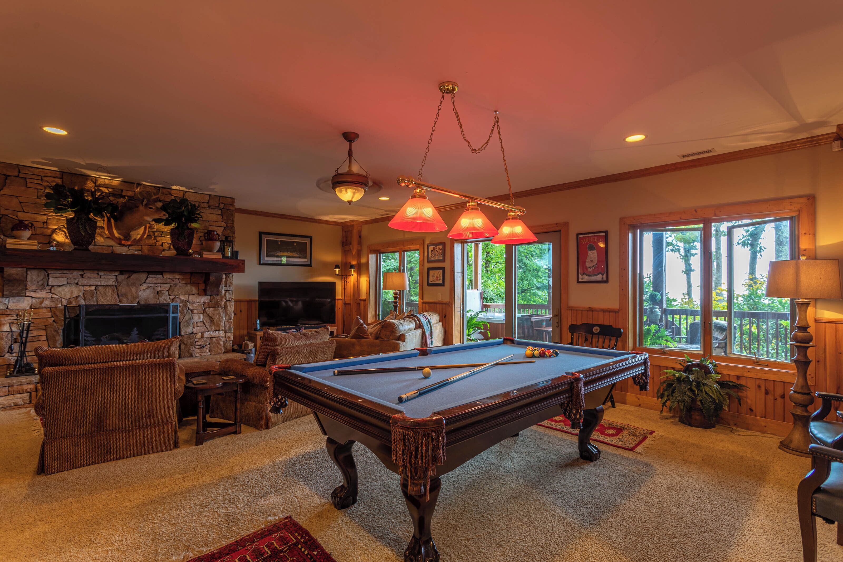 Un salon chaleureux avec une table de billard, des sièges confortables, une cheminée en pierre et de grandes fenêtres donnant sur l’extérieur.