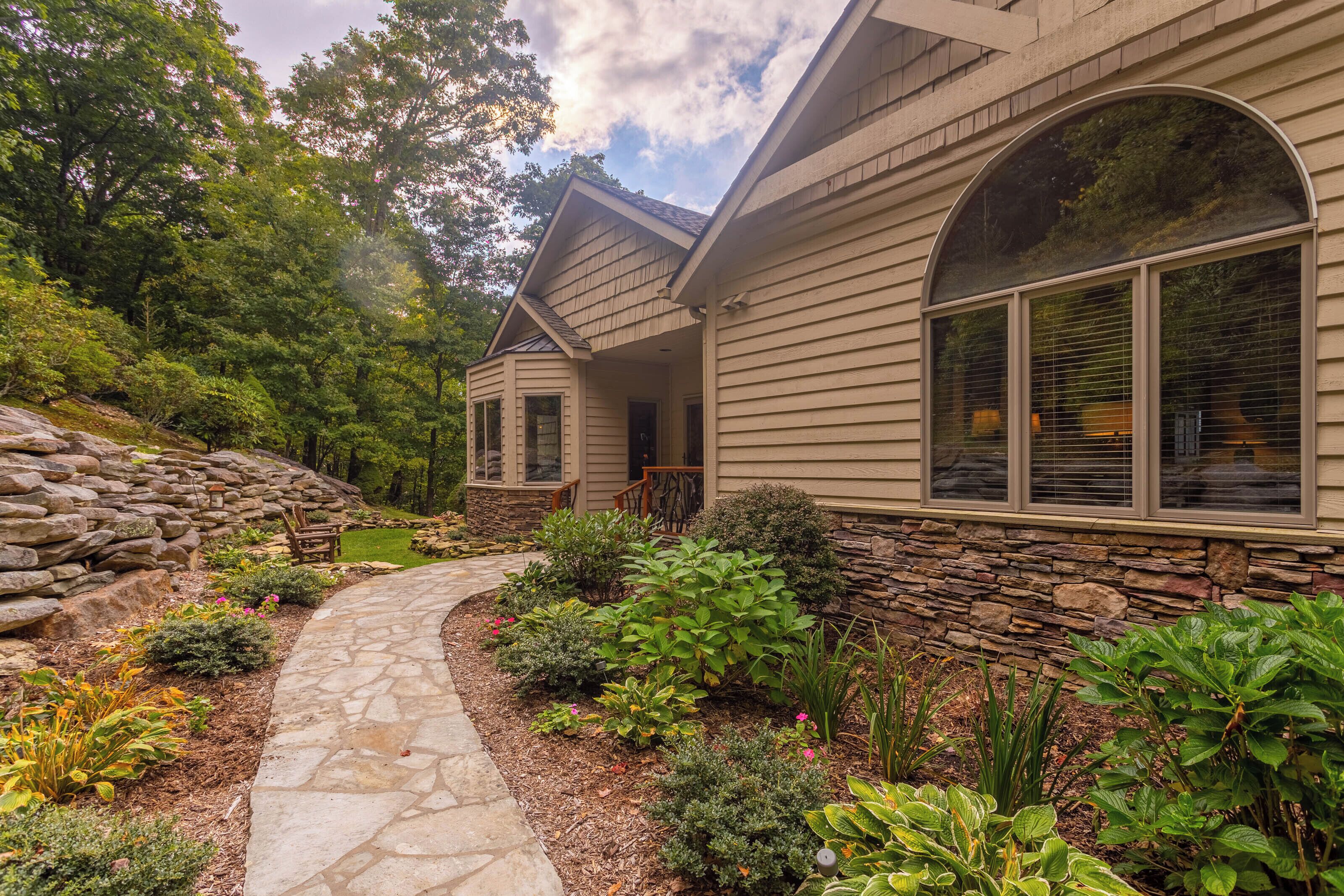 A house with a stone retaining wall, a stone pathway, and various shrubs and plants.