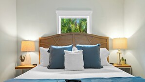 Een net opgemaakt bed met een blauwe sprei, een houten hoofdbord en meerdere kussens. Er staan twee nachtkastjes met lampen en boven het bed is een raam.