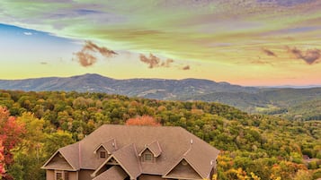 A house with a steeply pitched roof and a small porch, surrounded by dense forest and hills in the background.