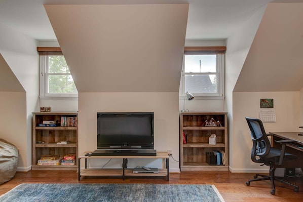 A loft-style room with a TV on a wooden stand, bookshelves, a desk with a chair, and a large window. - The Wander Inn (Blowing Rock)