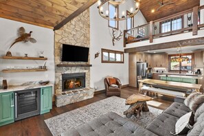 A spacious living area with a stone fireplace, a television mounted above it, and a dining area with a bench and table. - Eagle Rare Retreat at Eagles Nest (Banner Elk)