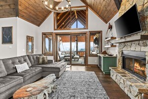 A spacious living area with a stone fireplace, a large flat-screen TV, and a view through a glass door to an outdoor deck. - Eagle Rare Retreat at Eagles Nest (Banner Elk)