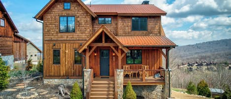 A two-story wooden cabin with a porch, stone foundation, and a view of a mountainous landscape.