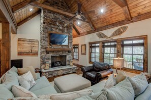 A cozy living room with a stone fireplace, leather sofa, and large windows. - Bluebird Cottage at Sweetgrass (Blowing Rock)