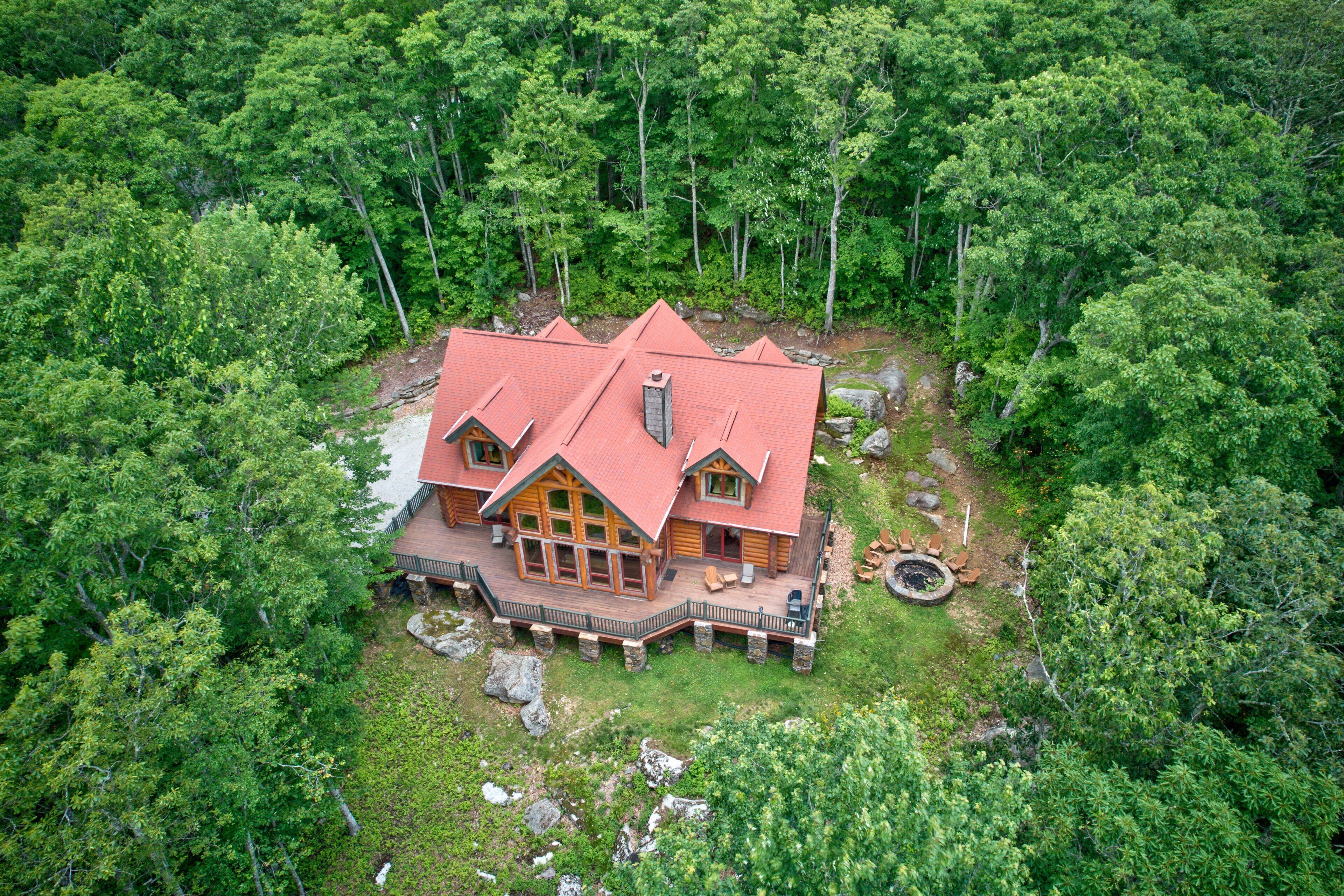 A large, log cabin-style house surrounded by dense forest, with a prominent red roof and a stone foundation.