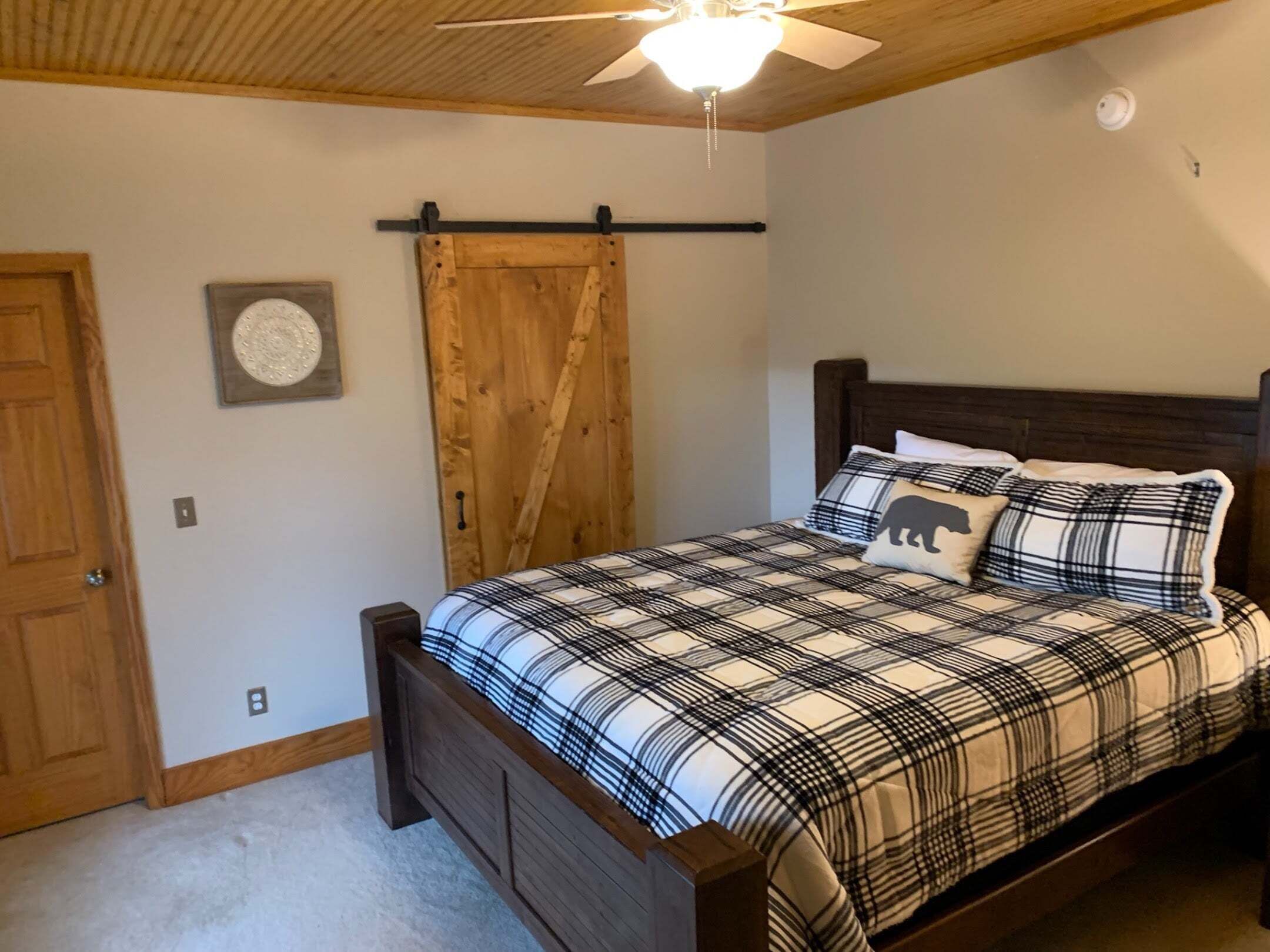 A bedroom with a bed, wooden headboard, a ceiling fan, a wooden door, and a wall-mounted light fixture.