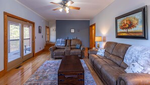 A living room with a ceiling fan, a brown leather sofa, a brown leather armchair, a wooden coffee table, and a painting of a tree. - Lazy Bear Inn (Sugar Grove)