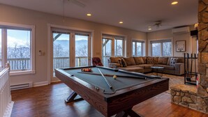 A pool table in a room with large windows, a ceiling fan, and a stone accent wall. - Blue Ridge Vista (Banner Elk)