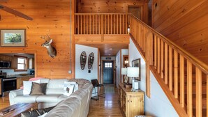 Interior de madera con un sofá, una cabeza de ciervo montada, una escalera y una zona de cocina.