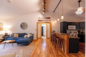 A modern living room with a blue sofa, wooden flooring, and a kitchen with dark cabinets and a white microwave.