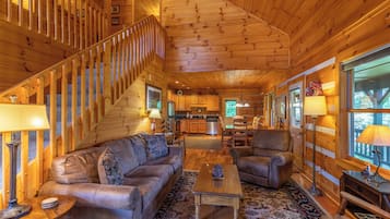 A log cabin interior with a living room, a staircase, a kitchen, and large windows.