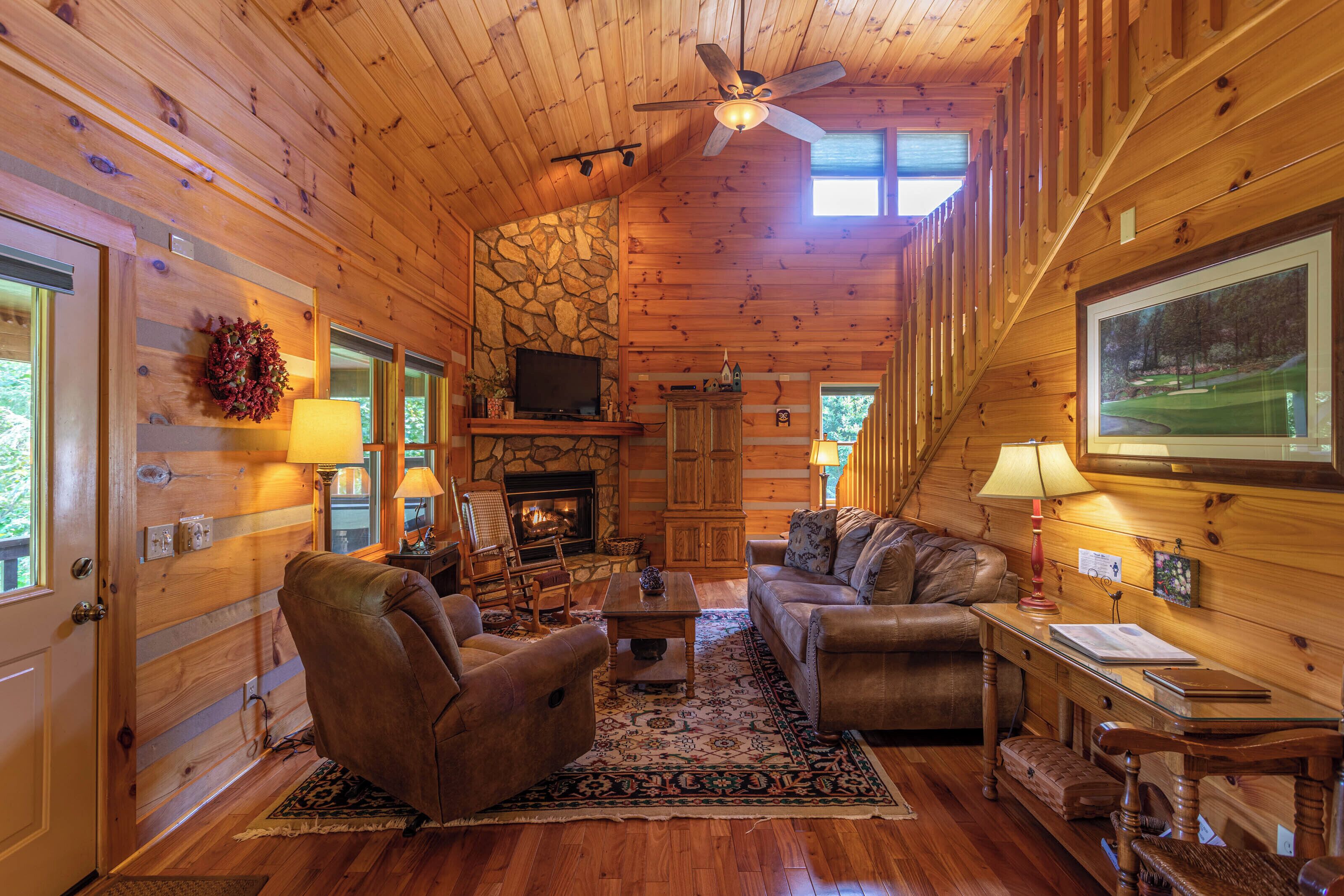 A cozy cabin interior with a fireplace, wooden walls, a ceiling fan, and a large window.