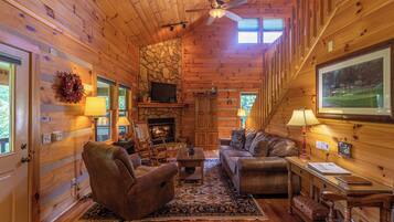 A cozy cabin interior with a fireplace, wooden walls, a ceiling fan, and a large window.