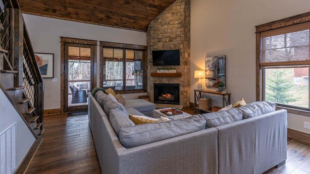 A spacious living room with a stone fireplace, a large gray sectional sofa, and a wooden staircase. - Overlook Nook at Eagles Nest (Elk Park)