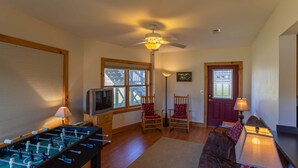 A room with a foosball table, a television, two rocking chairs, a door with a window, and a ceiling fan. - River House at Meadow Landing (Fleetwood)