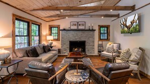 A cozy living room with a fireplace, wooden beams, a sofa, leather chairs, and a television mounted on the wall. - Moosey Creek Lodge (Banner Elk)