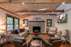 A cozy living room with a fireplace, wooden beams, a sofa, leather chairs, and a television mounted on the wall.