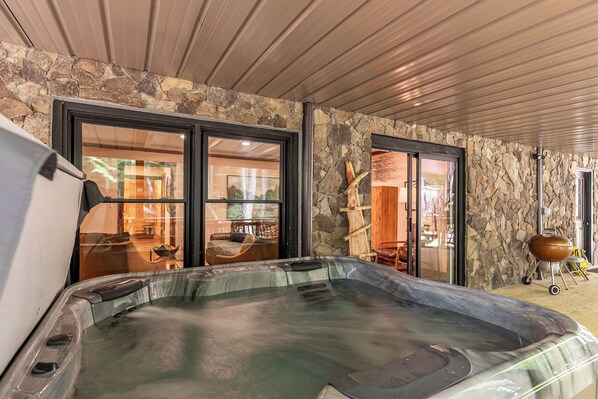 A hot tub in a covered outdoor area with stone walls and large sliding glass doors leading to an interior space.