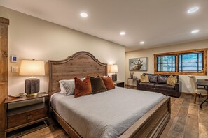 A bedroom with a large bed, two bedside tables with lamps, a wooden headboard, a brown leather sofa, a small table, and a window with blinds.