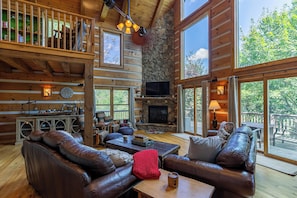 A log cabin interior with a stone fireplace, leather sofas, a wooden coffee table, and large windows offering a view of the outdoors. - Cari's Rivers Edge (Boone)