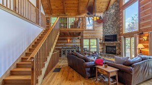 Ein geräumiges Blockhaus-Interieur mit einer Holztreppe, Ledersofas, einem Kamin und großen Fenstern.