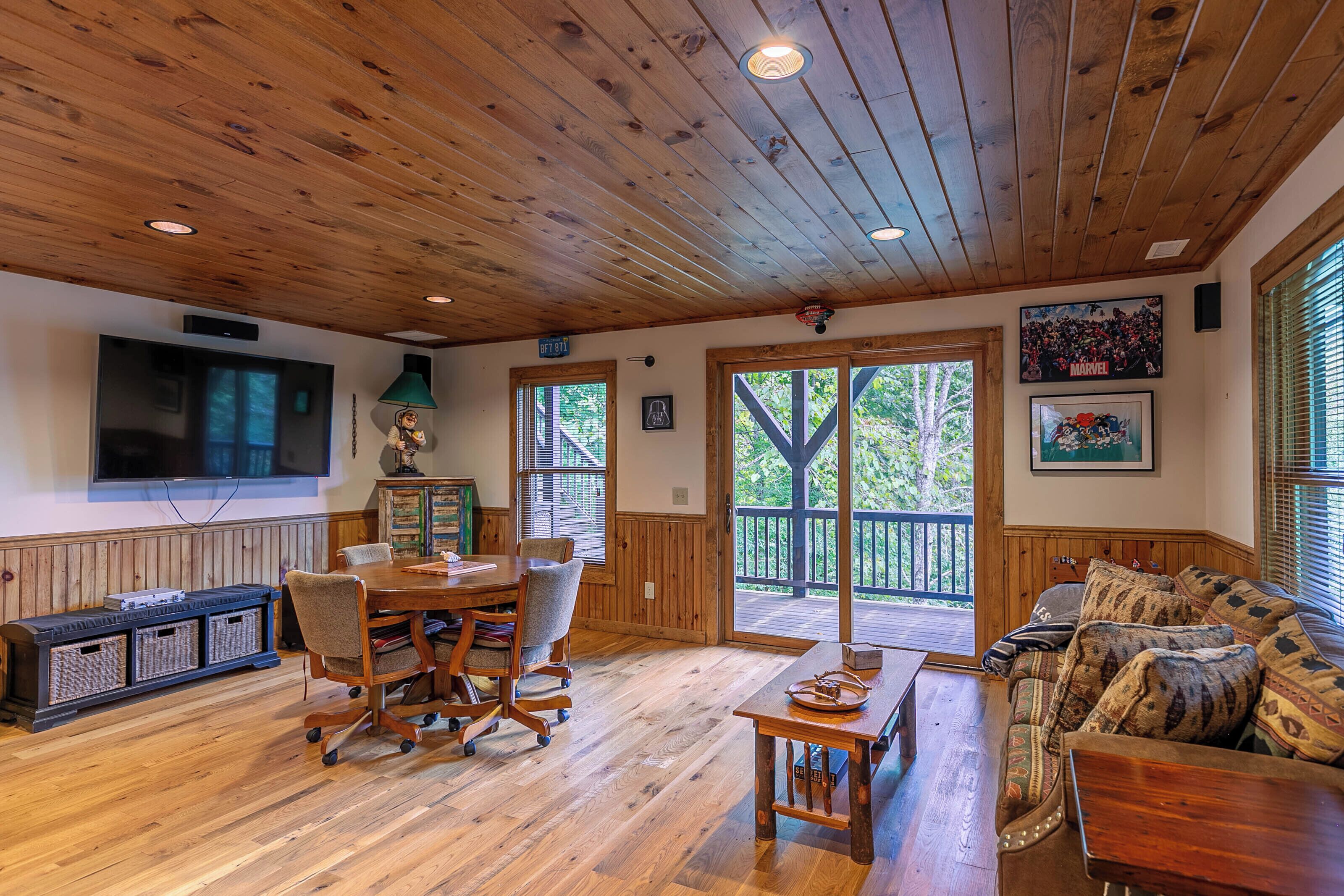 A wooden-floored living room with a dining area, a television mounted on the wall, and a large window with a view of trees.