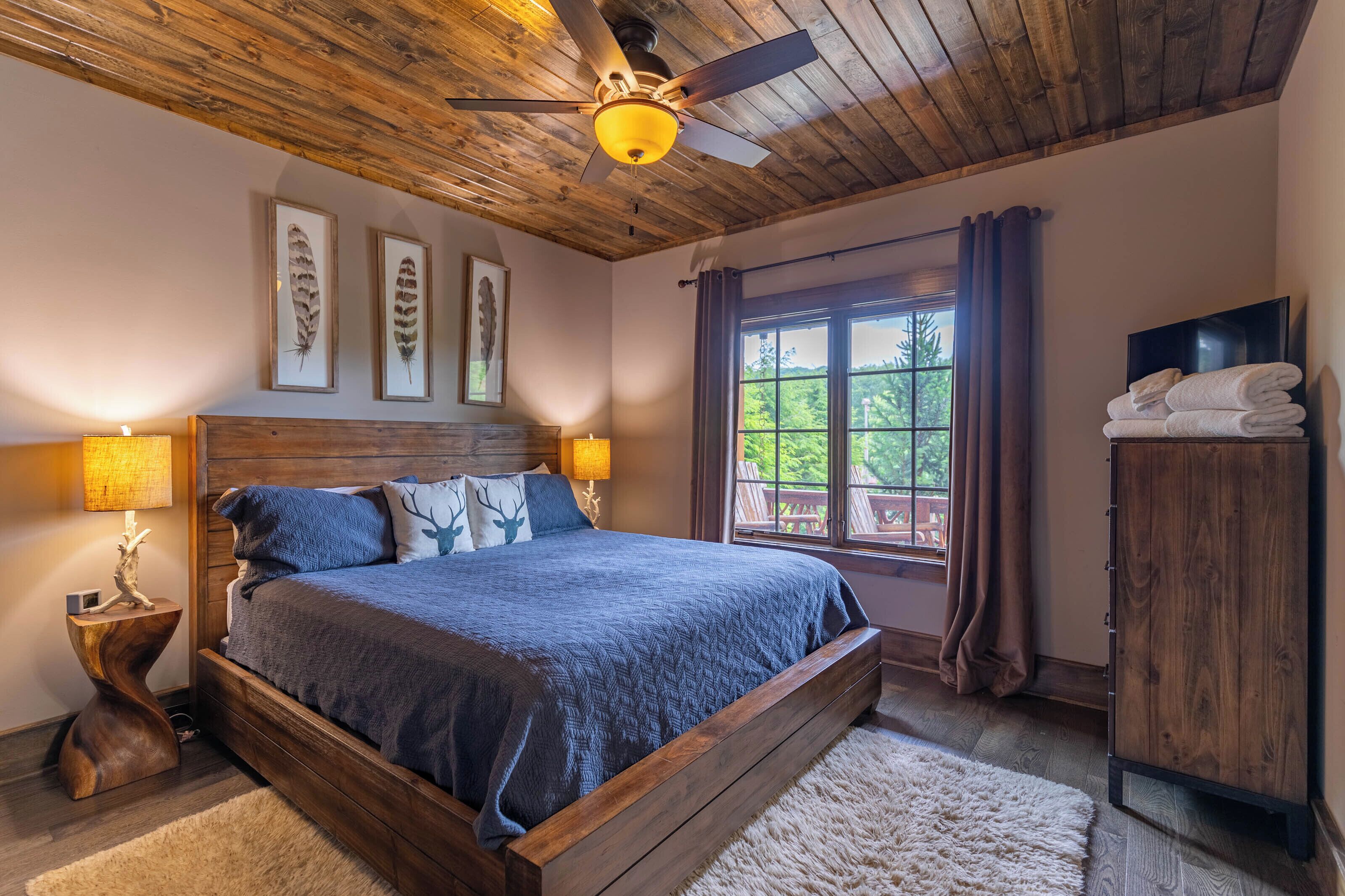 Une chambre avec un plafond en bois, un lit recouvert d’une couverture bleue, deux lampes fixées au mur, une fenêtre avec des rideaux et un téléviseur posé sur une commode en bois.