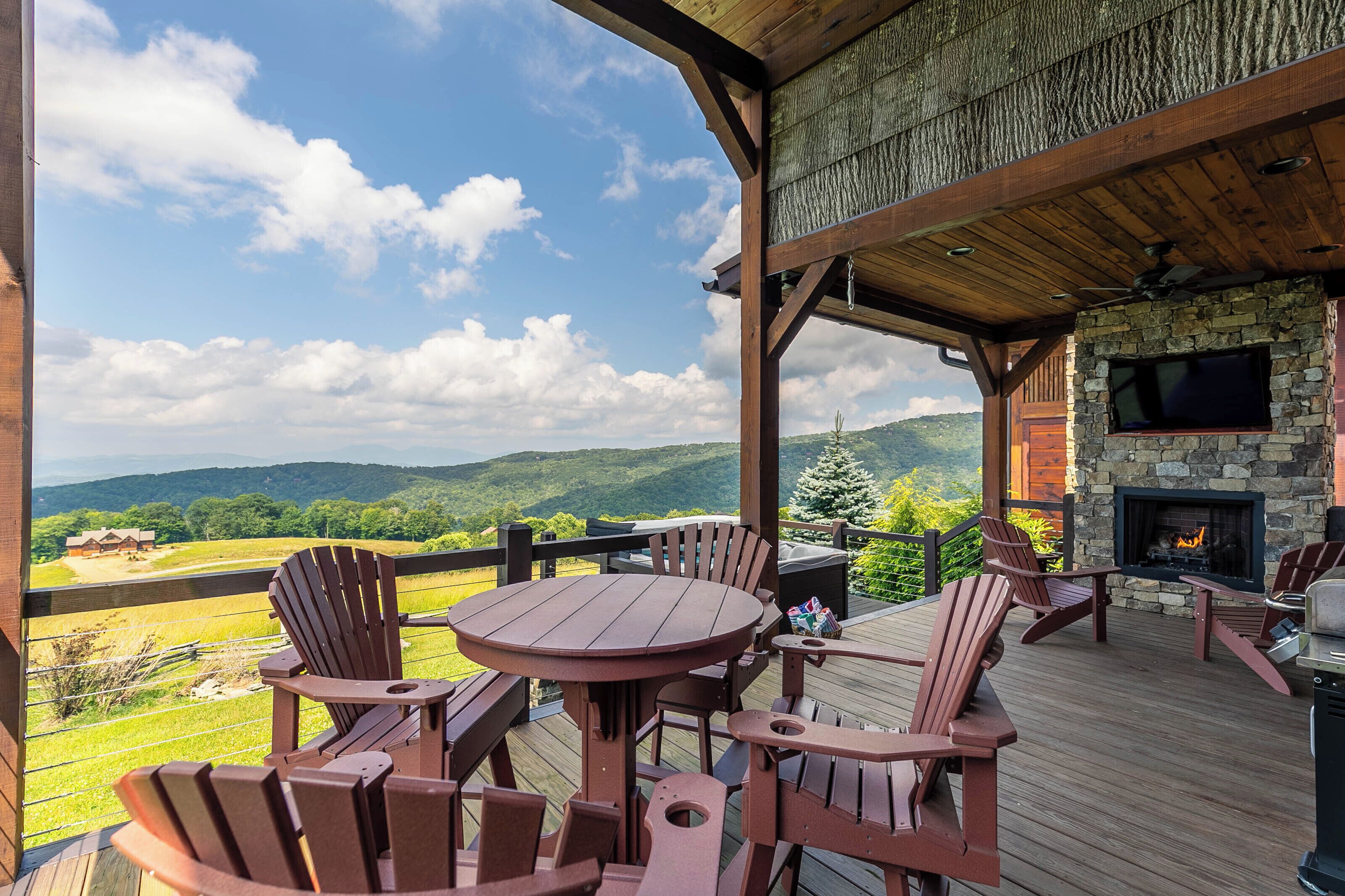 Una terraza de madera con sillas Adirondack y una mesa redonda, con vistas a un paisaje pintoresco con montañas y una casa a lo lejos.
