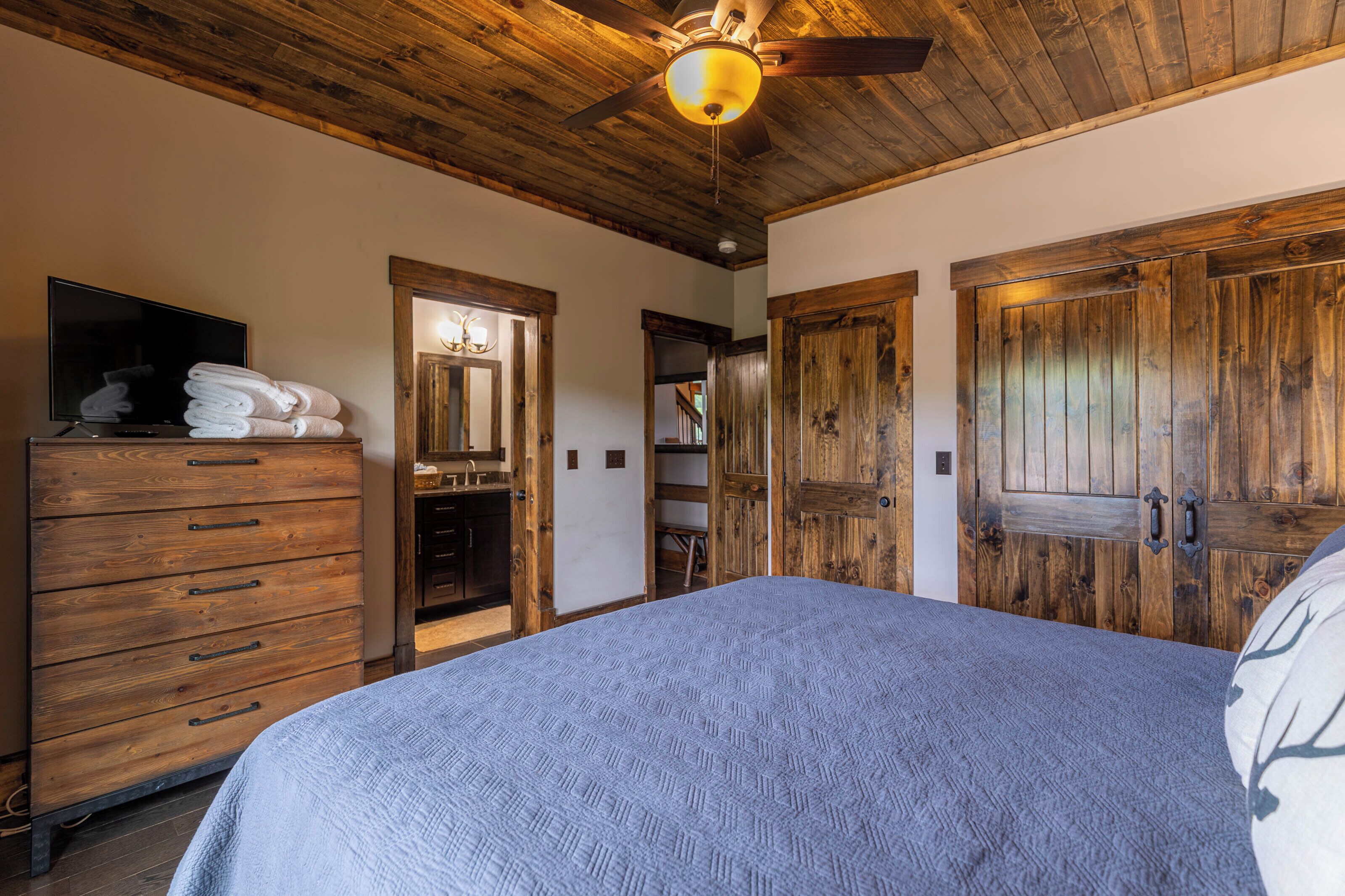 A bedroom with a wooden ceiling, a bed with a textured blanket, a wooden dresser with a TV, and a wooden wardrobe.