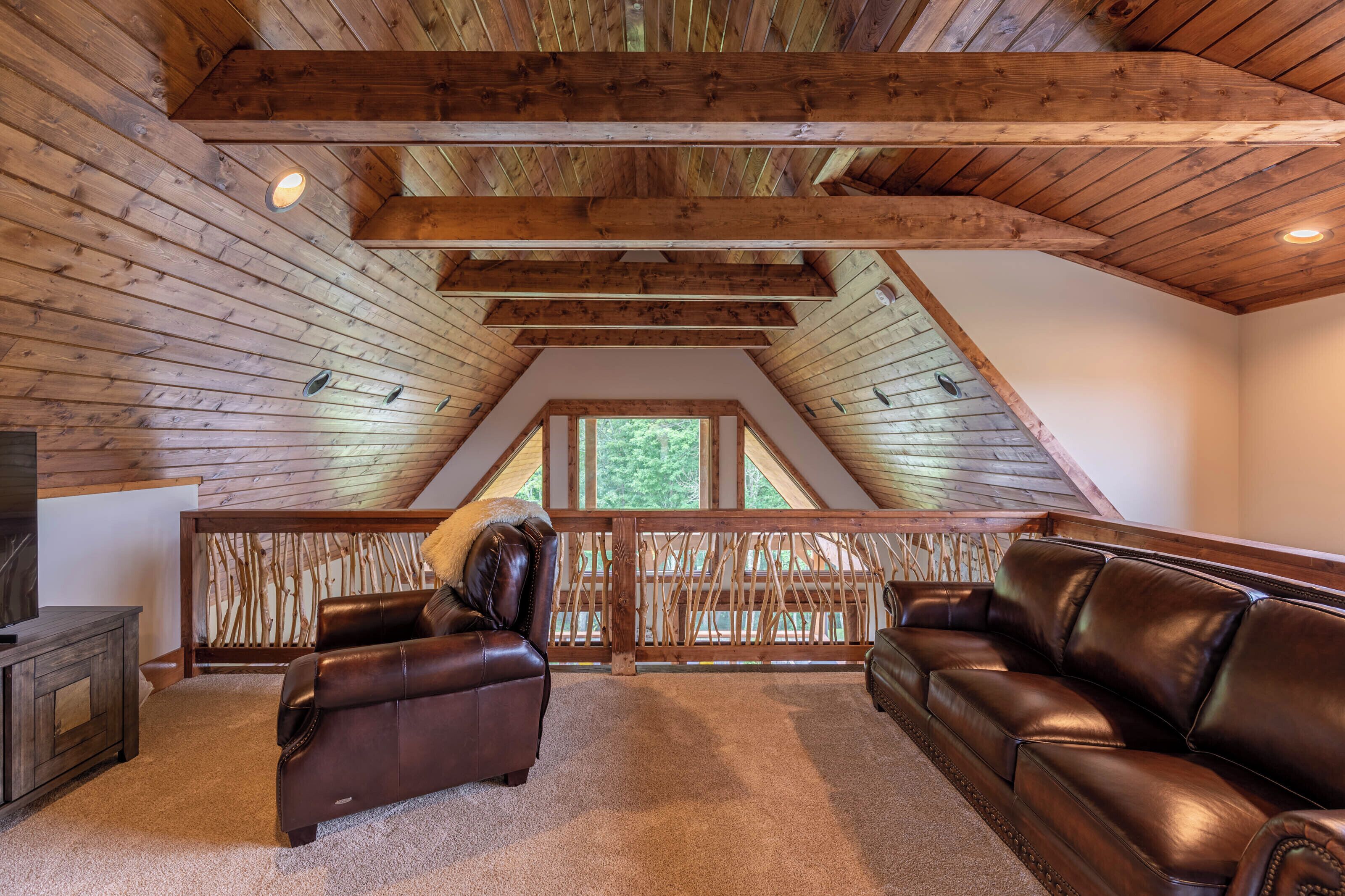 Een loft-achtige kamer met zichtbare houten balken, een schuine kap en een uitzicht door een groot raam. Er staan twee leren banken en een houten console tafel.