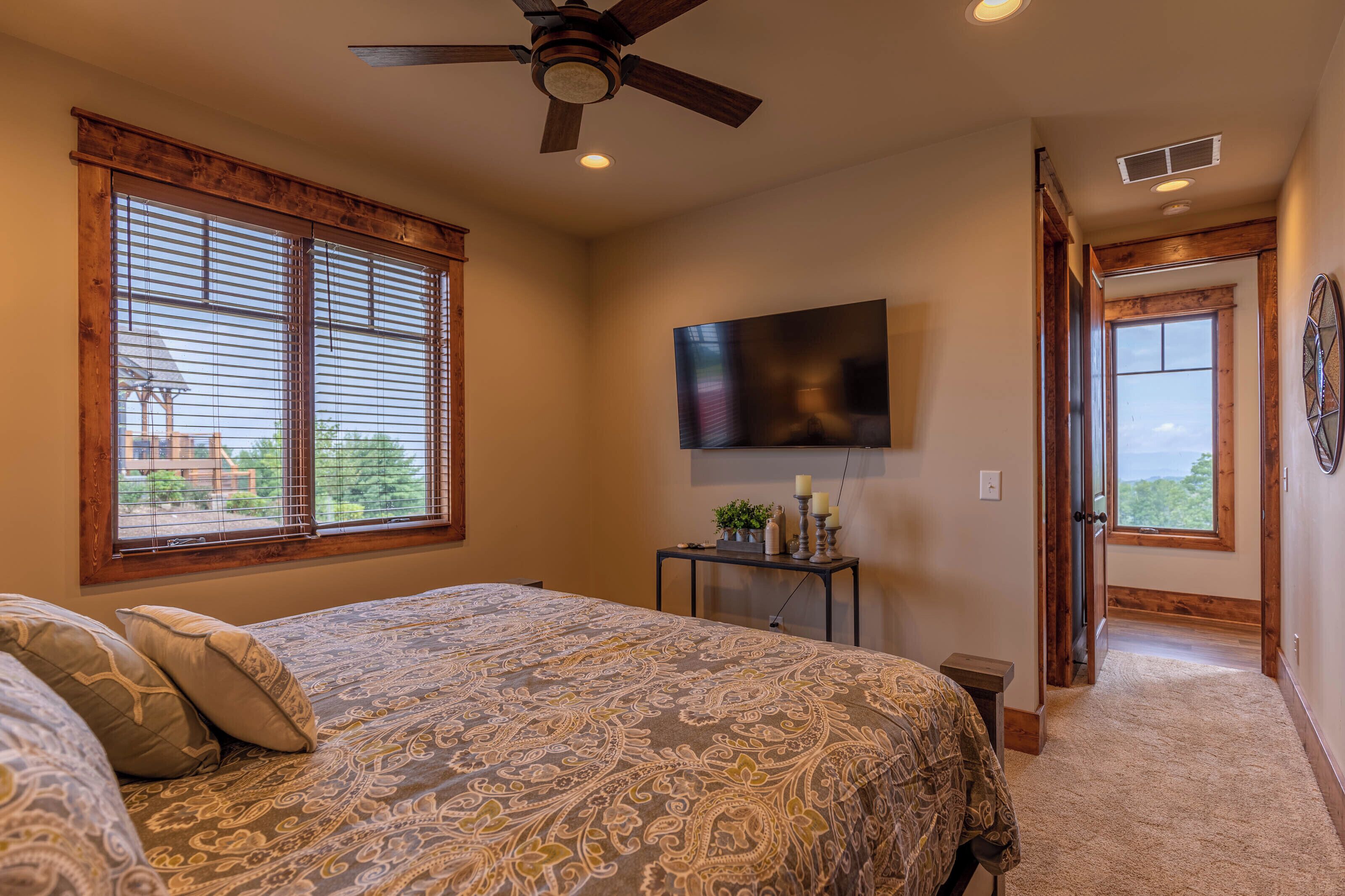 A bedroom with a large bed, a ceiling fan, a TV mounted on the wall, and a view of the outdoors through the windows.