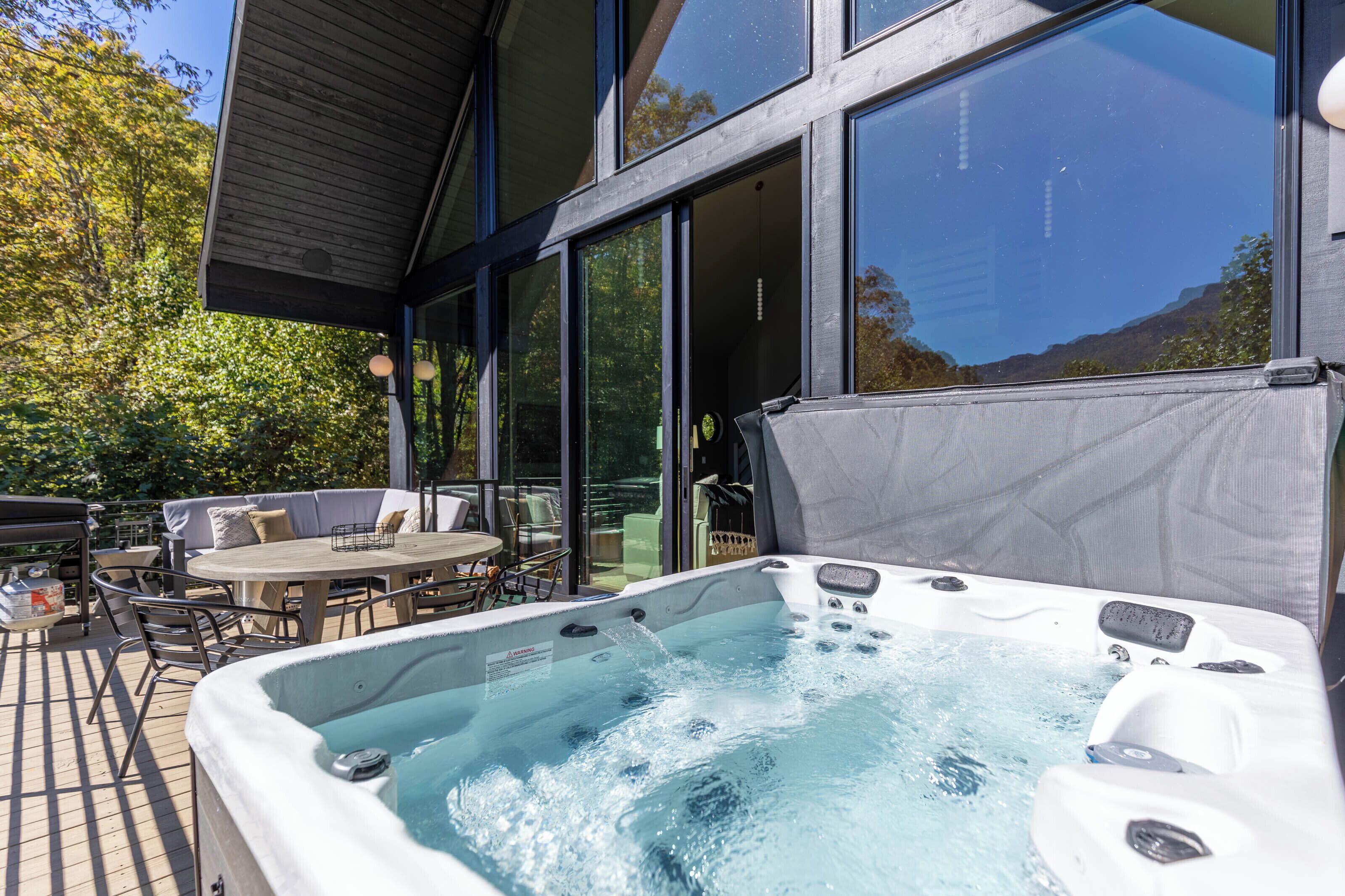 Un bain à remous doté d’une terrasse en bois et de mobilier d’extérieur, entouré de grandes fenêtres et offrant une vue sur les arbres.