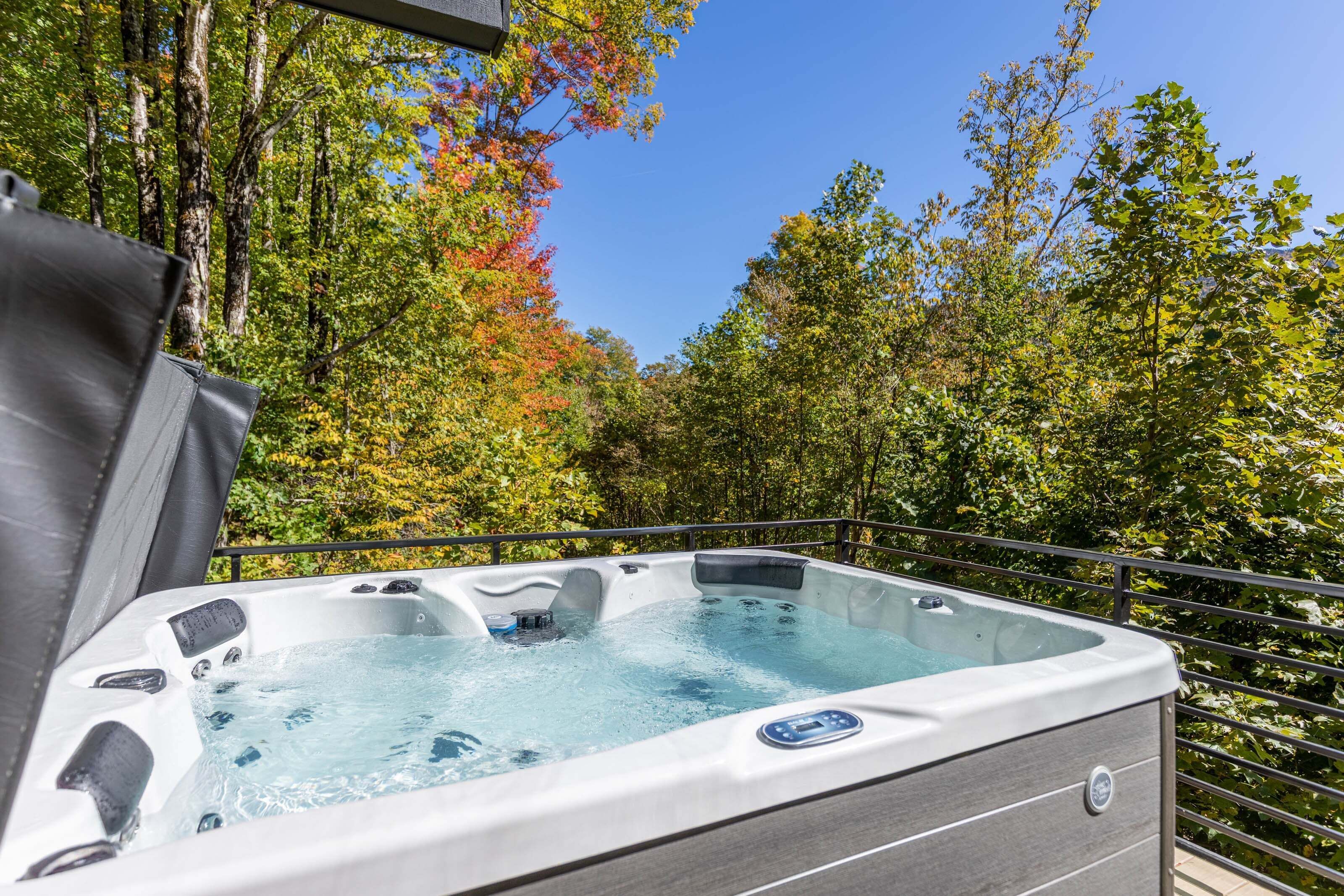 A hot tub with a control panel, surrounded by a metal railing, set against a backdrop of trees and a clear sky.