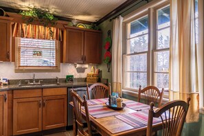 A kitchen with wooden cabinets, a window with curtains, a dining table with chairs, and a window view.