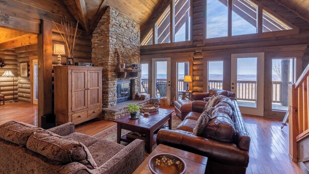 A log cabin interior with a stone fireplace, leather sofas, and large windows offering a view. - Highland Mist (Banner Elk)