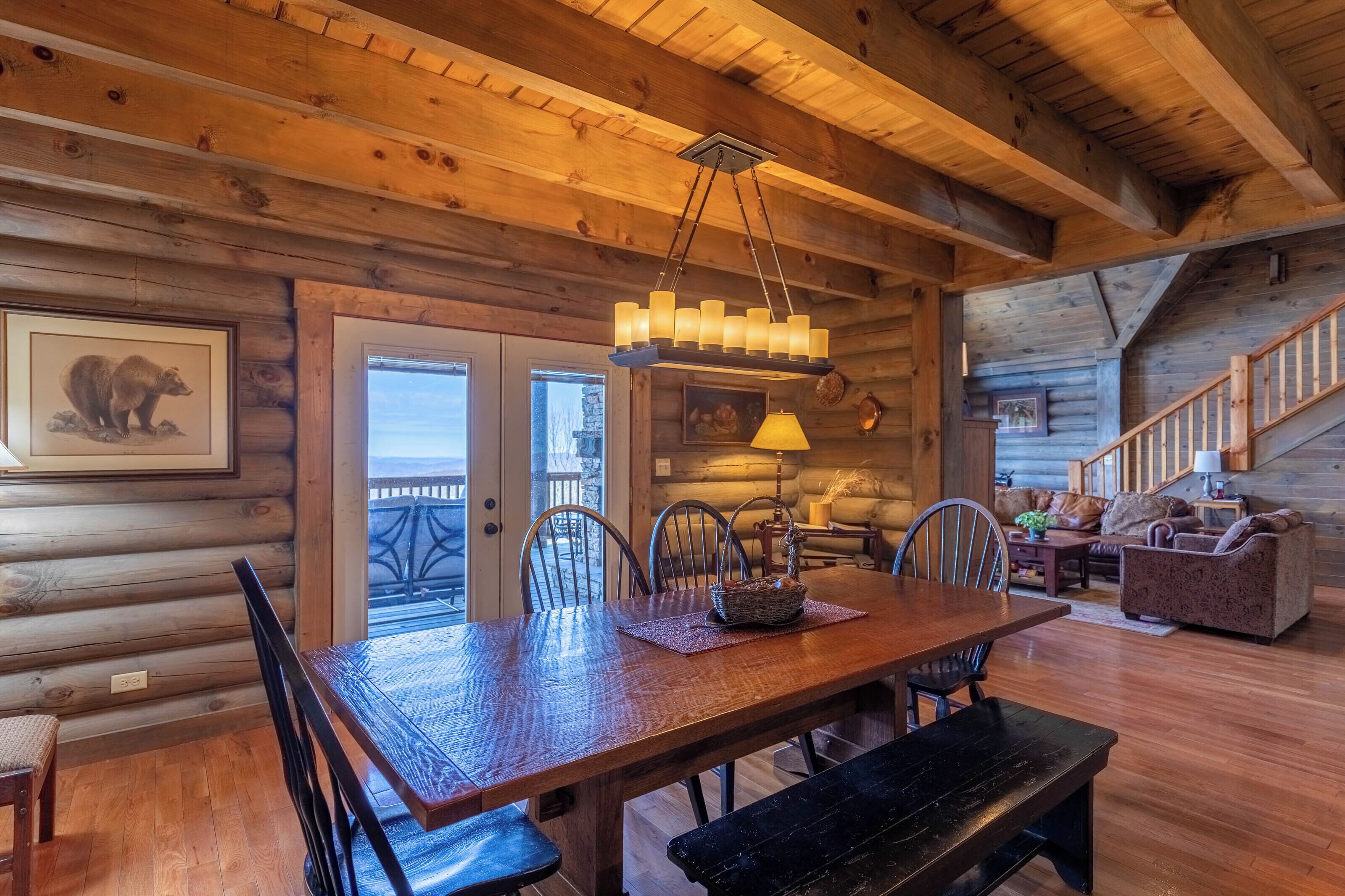 A cozy log cabin interior with a wooden dining table, chairs, a bench, a staircase, and a living area with a sofa and a lamp.