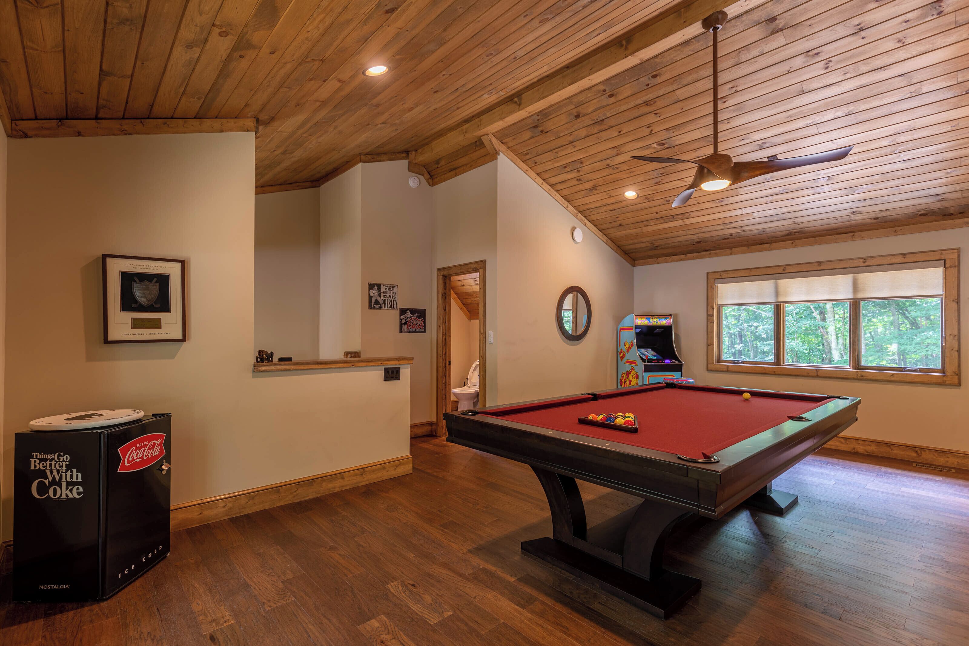 A pool table in a room with wooden floors and ceiling, a ceiling fan, and a window with a view of trees.