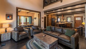 Ampla sala de estar com um sofá grande, poltronas e uma mesa de centro. Cozinha com armários de madeira e área de jantar com mesa e cadeiras são visíveis.