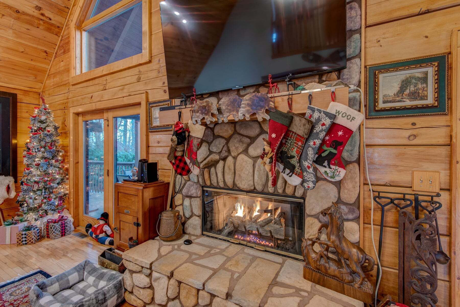A cozy log cabin interior with a stone fireplace, Christmas stockings, a decorated tree, and a television.