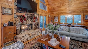 A cozy cabin interior with a stone fireplace, wooden walls, a sofa, and a dining area. - Blueberry Hills 3BR With hot Tub, pet Friendly, Outdoor Fireplace (Boone)