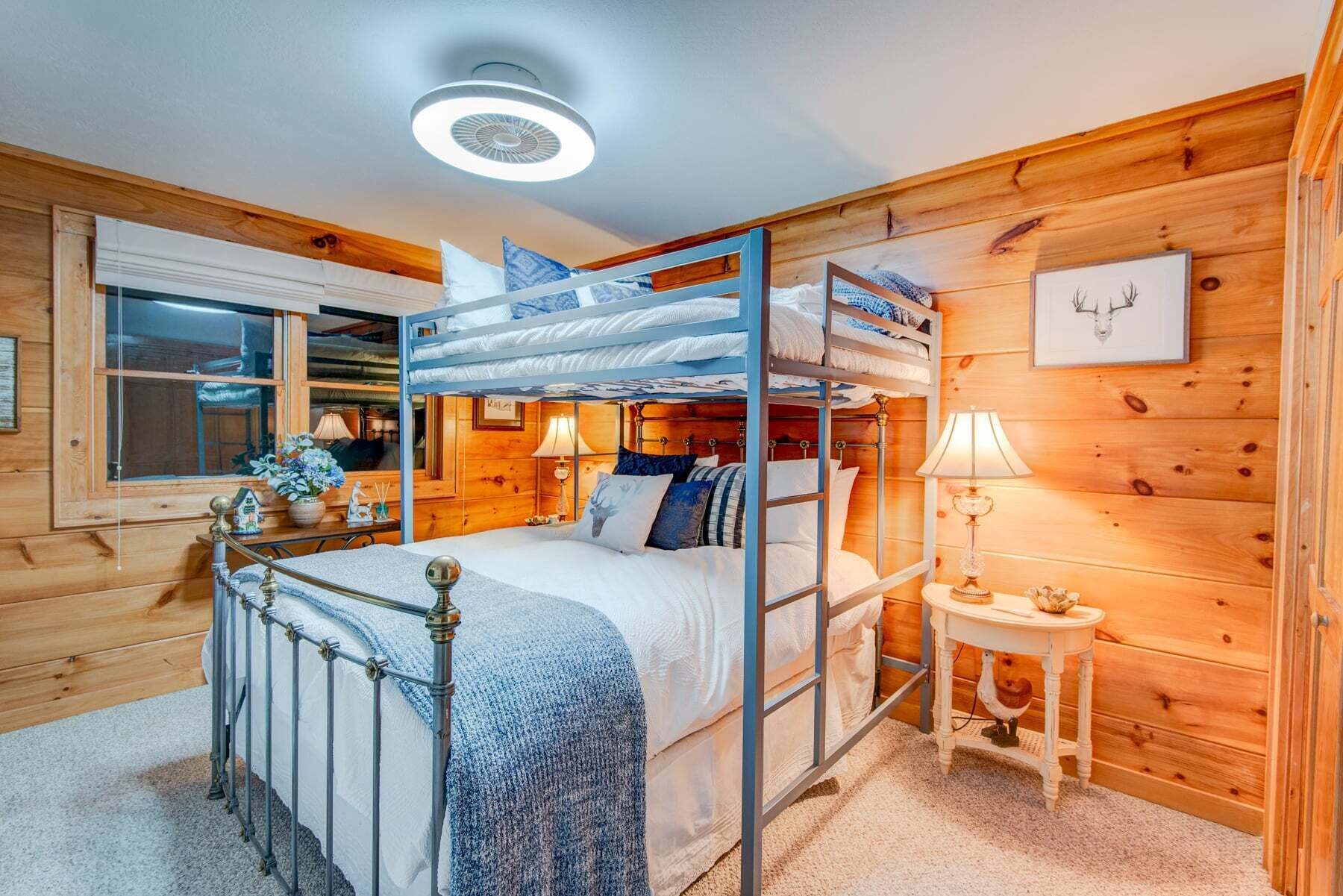A bunk bed room with a window, bedside table, lamp, and a framed picture on the wall.