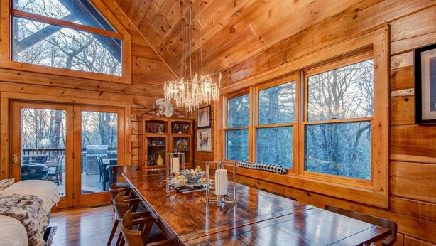 A wooden cabin interior with a large dining table, chairs, a chandelier, and large windows offering views of trees. - Blueberry Hills 3BR With hot Tub, pet Friendly, Outdoor Fireplace (Boone)