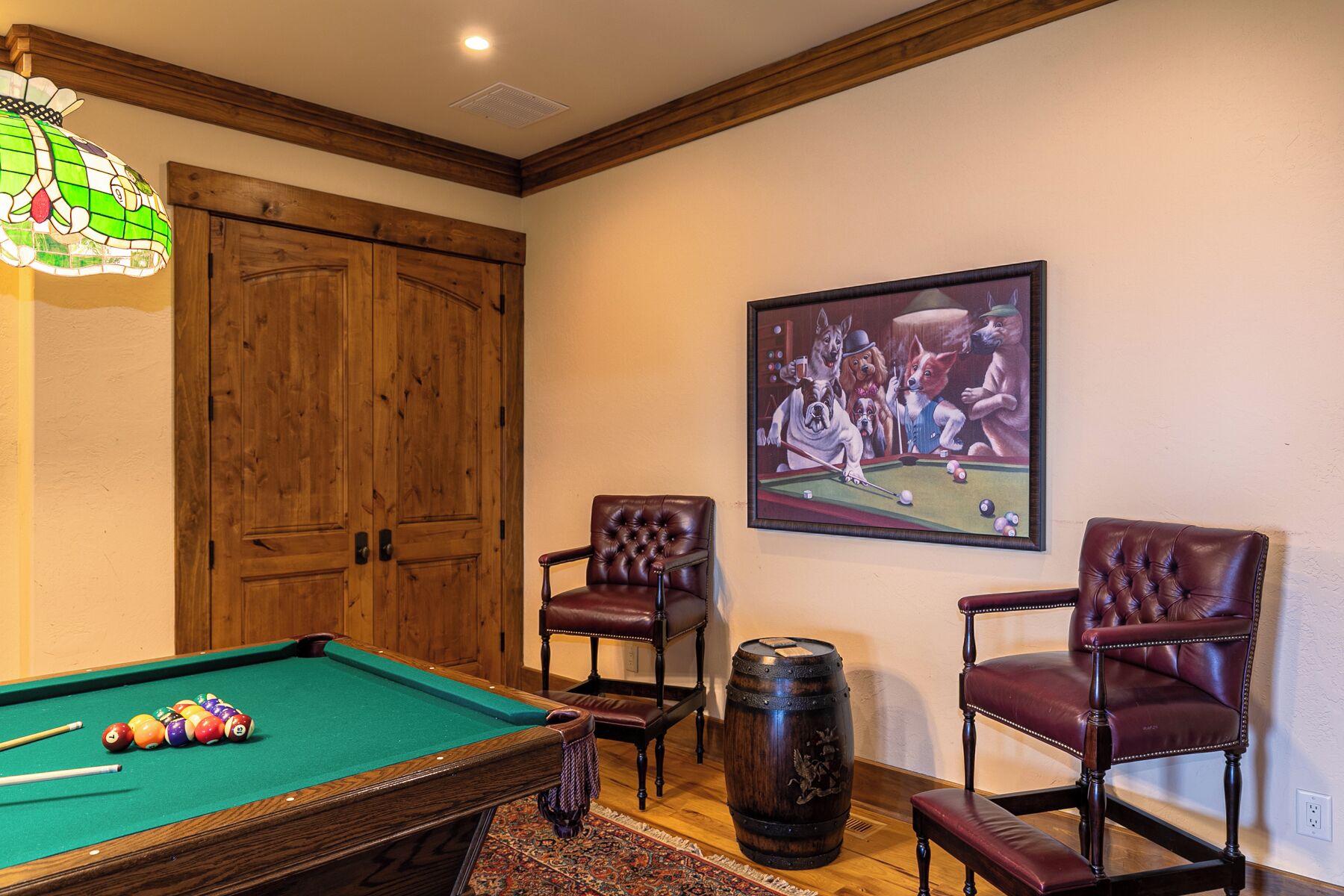 A pool table with balls, two leather chairs, a wooden door, and a painting of dogs playing pool.