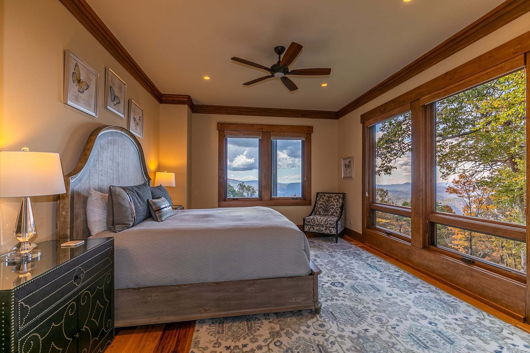 A bedroom with a bed, a chair, a desk, a lamp, a ceiling fan, and large windows with a view.