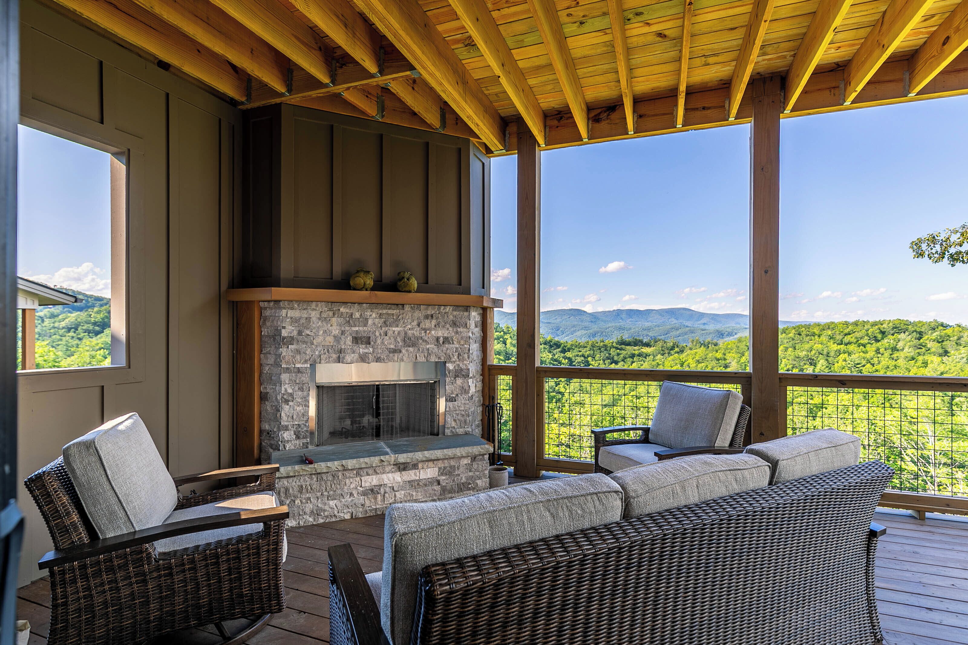 A covered outdoor seating area with wicker furniture, a stone fireplace, and a view of a mountainous landscape.