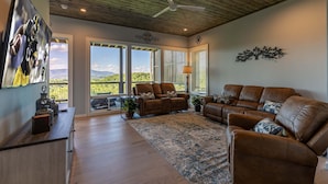 A spacious living room with a large brown leather sofa, a coffee table, and a TV on a wooden stand. There are large windows with a view of the outdoors. - Blue Ridge Sanctuary at Blue Ridge Mountain Club (Lenoir)