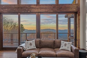 A room with a large window, a brown leather sofa, and a view of mountains. - The Tipsy Bear at Eagles Nest (Elk Park)