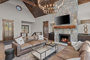 A spacious living room with a stone fireplace, leather sofas, a glass coffee table, and a mounted TV.
